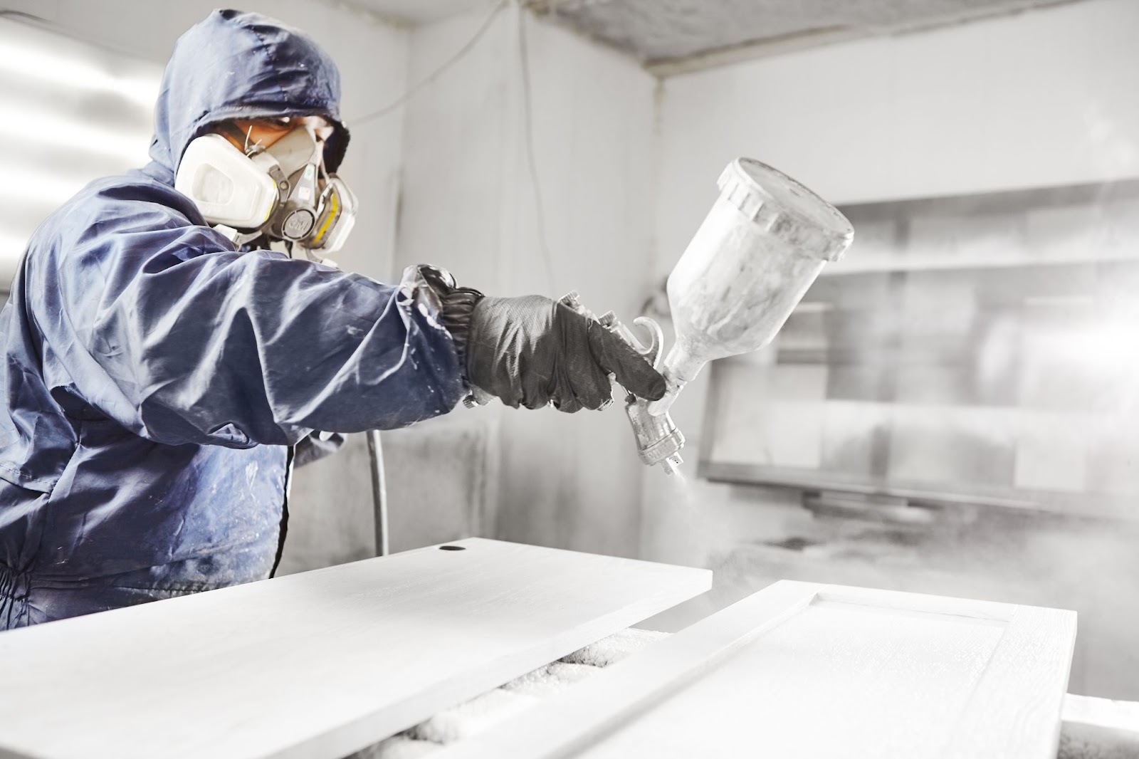 A masked man in a blue jacket applies white paint to cabinets, demonstrating expertise in professional cabinet finishing.