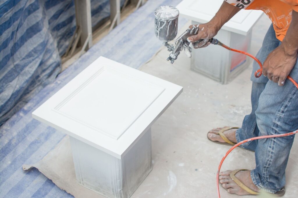 A man expertly uses a spray gun to paint a white table, showcasing professional cabinet painting skills.