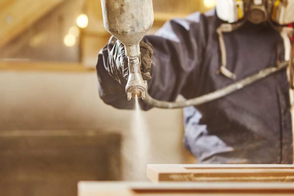 A professional cabinet painter in a blue jacket and safety glasses uses a spray gun to apply finish to wooden surfaces.