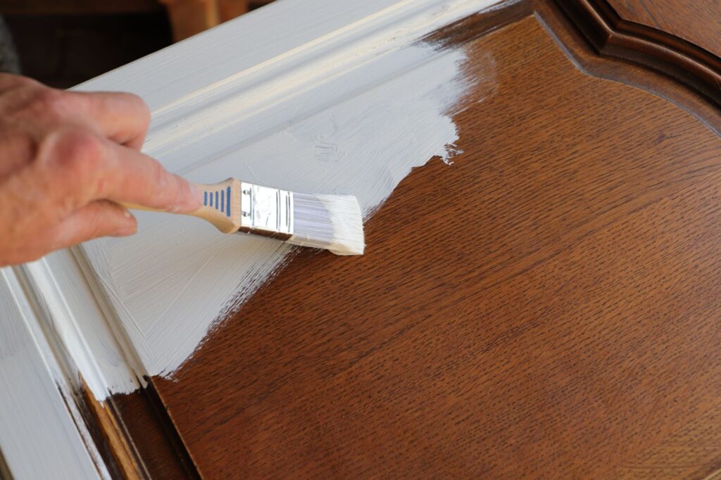 A person applies white paint to a wooden door, preparing it for a fresh look in a kitchen cabinet renovation.