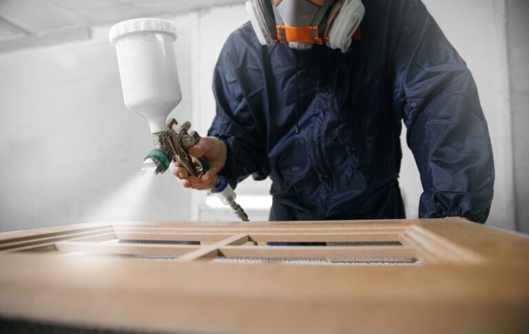 A man in a protective suit sprays a wooden door, showcasing cabinet painting and home improvement techniques.