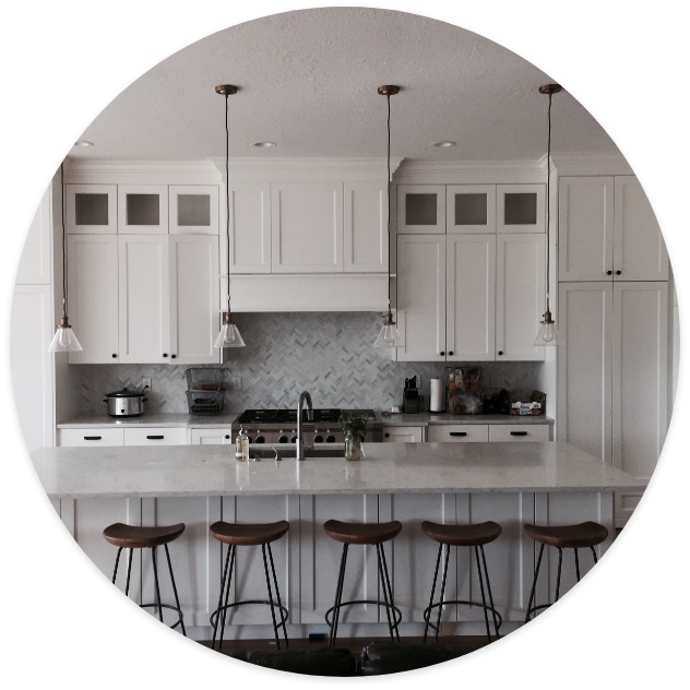 Modern white kitchen with a large island, bar stools, and pendant lighting, featuring a herringbone tile backsplash and stainless steel appliances