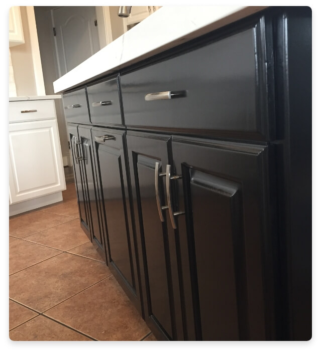 Close-up view of a kitchen island with black refinished cabinets, sleek silver handles, and a white countertop, showcasing a modern contrast with tiled flooring.