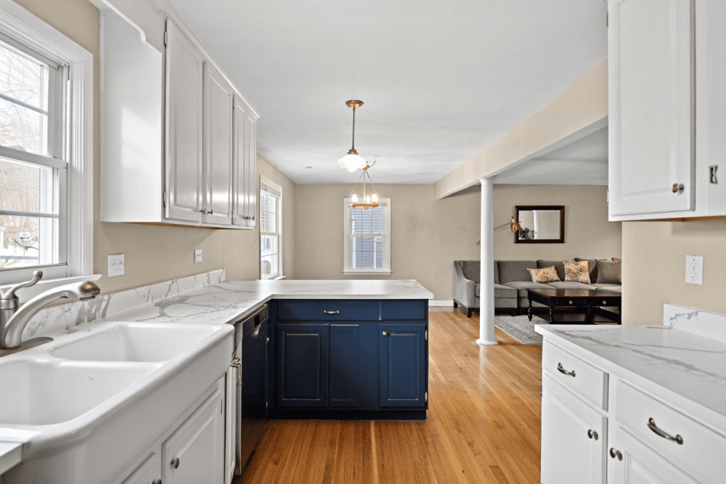 A photorealistic interior design photograph of a bright, newly renovated transitional kitchen. The perimeter kitchen cabinets are painted a clean, warm white with shaker-style doors. The large central island is painted a contrasting deep navy blue, making it the elegant focal point of the room. The kitchen flows into a living area in the background which has neutral tones, showing that the Kitchen Cabinets Match the Rest of Your Home's aesthetic. The finish on the painted cabinets looks flawless and professional. Natural light streams in from a large window over the sink.