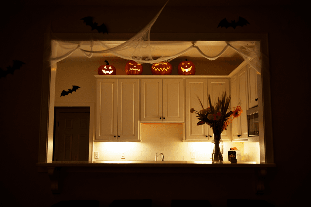 A wide view of a white kitchen showing how to decorate your kitchen by utilizing the space above upper cabinets with faux cobwebs and ceramic jack-o-lanterns.