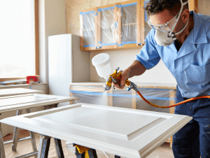 Pro spraying satin white 2K polyurethane on kitchen cabinet doors; kitchen masked with plastic and blue painter’s tape.