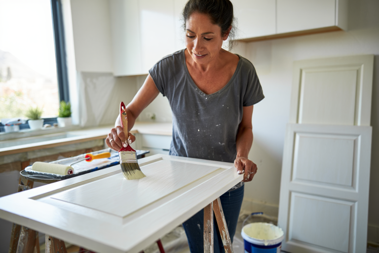 Homeowner painting kitchen cabinets with brush, showing visible stroke marks on white cabinet door panel