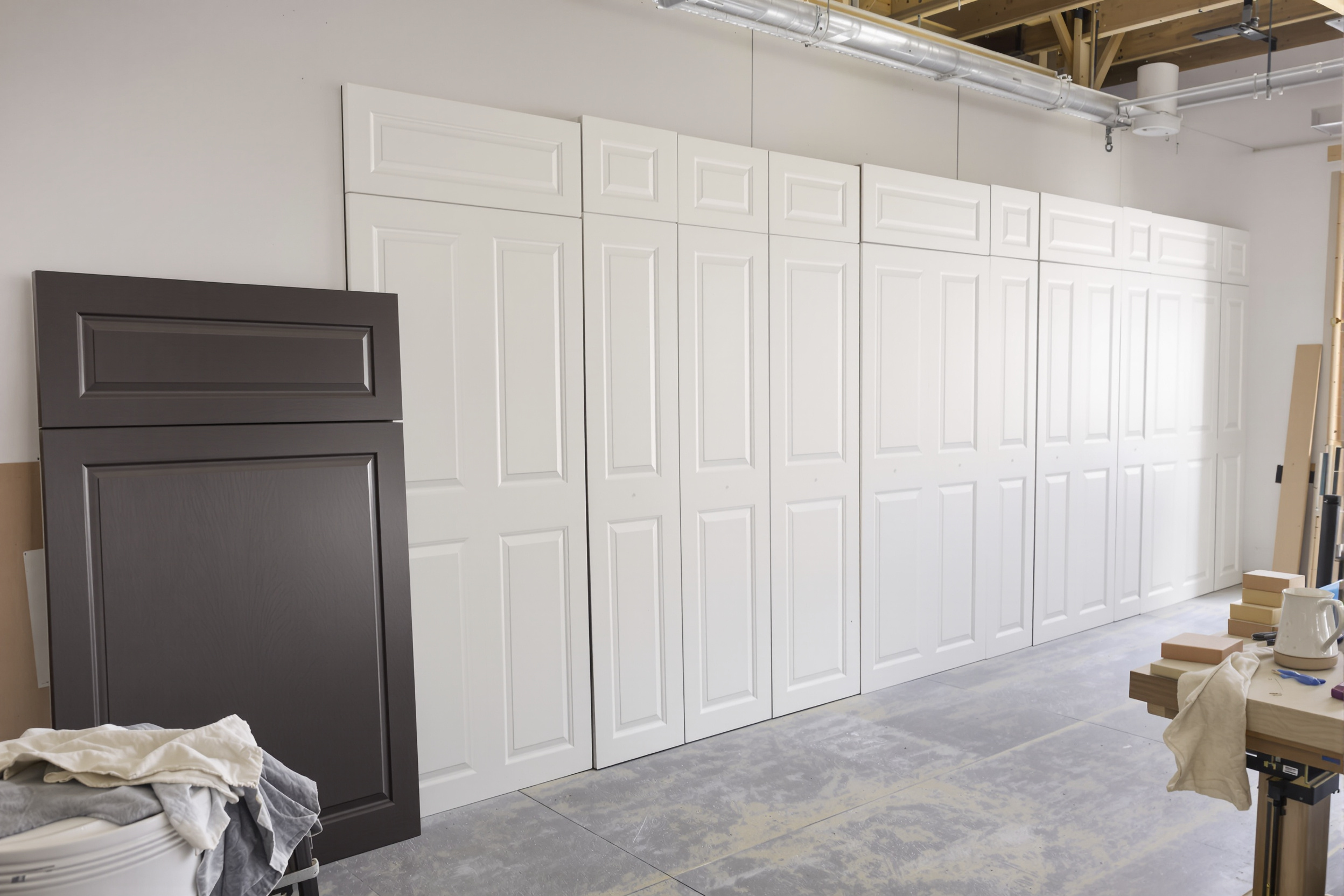 Cabinet doors from a dark stained kitchen coated in white primer beside an unprimed espresso door in an off-site finishing shop