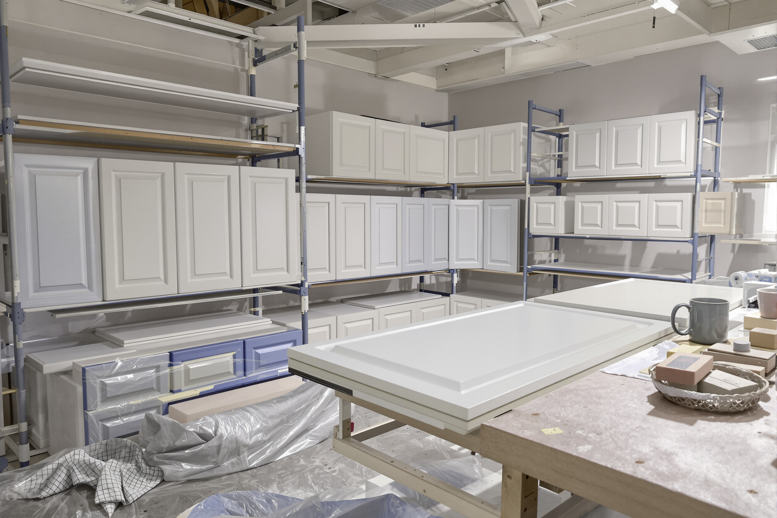 Previously painted cabinet doors sanded and coated in white primer in an off-site finishing shop beside drawer fronts still in their old blue paint